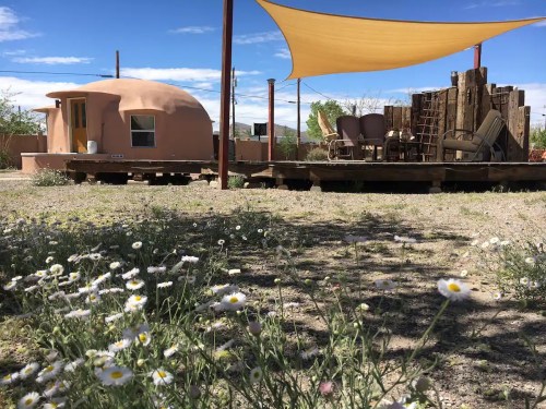 A desert scene featuring a round adobe structure, a wooden deck with chairs, and wildflowers in the foreground.