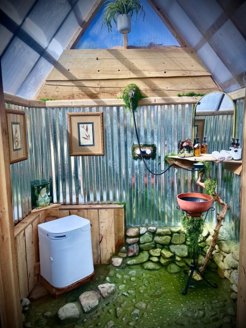 A unique indoor space featuring a compost toilet, stone floor, and greenery, with framed art on the walls.