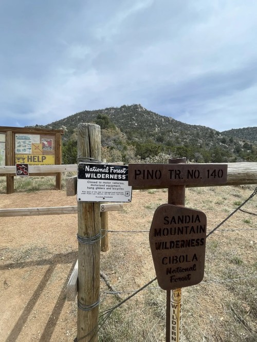Signpost marking the Pino Trail No. 140 in the Sandia Mountain Wilderness, with a mountain in the background.