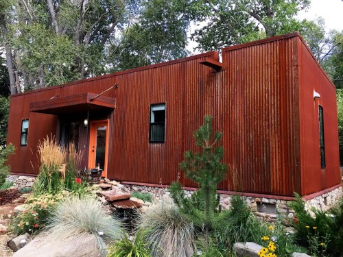 A modern, rust-colored metal house surrounded by greenery and colorful flowers, featuring a welcoming front entrance.