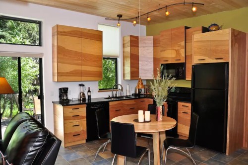 Modern kitchen with wooden cabinets, black appliances, a round dining table, and large windows overlooking greenery.