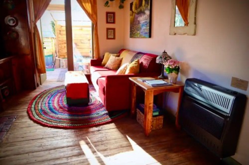Cozy living space with a red sofa, colorful rug, wooden table, and warm lighting from curtains and a lamp.