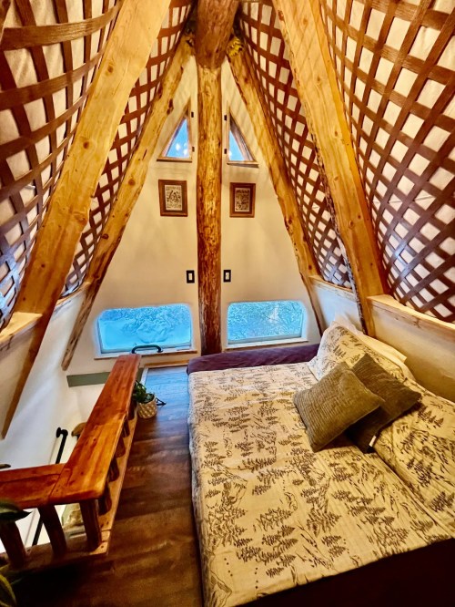 Cozy attic bedroom with wooden beams, a bed, and large windows overlooking nature. Warm, inviting atmosphere.