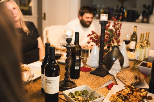 A cozy dinner table with wine bottles, food, and candles, featuring guests enjoying a meal in a warm setting.