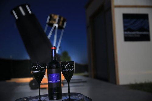 A bottle of wine and two glasses in front of a large telescope at night, with a logo in the background.