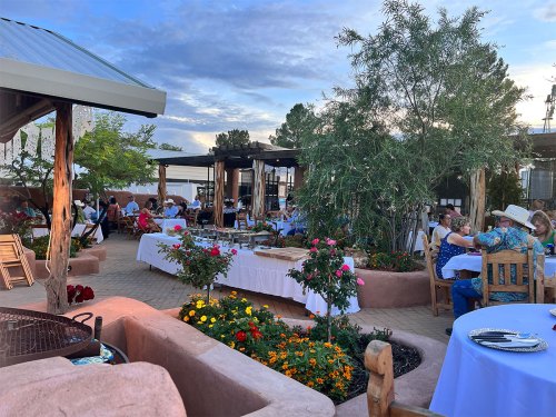 Outdoor dining area with tables, flowers, and guests enjoying a meal under a blue sky with scattered clouds.