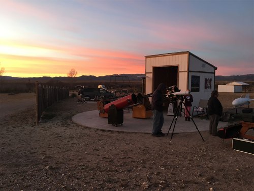 A sunset scene at an observatory with telescopes and people setting up for stargazing. Mountains in the background.