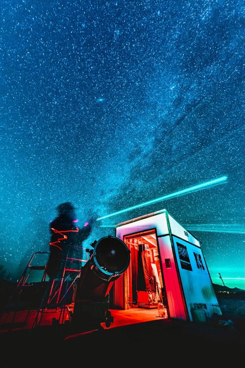 A person observes the starry sky with a telescope, illuminated by red lights, near a small observatory building.