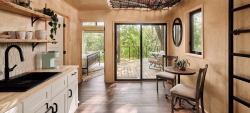 Cozy interior of a modern cabin with a kitchen, dining area, and large windows overlooking a deck and greenery.