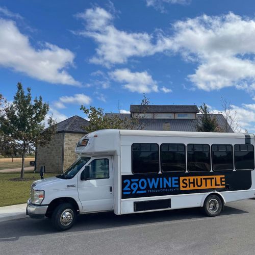 A white shuttle bus with "290 Wine Shuttle" branding parked near a building under a partly cloudy sky.