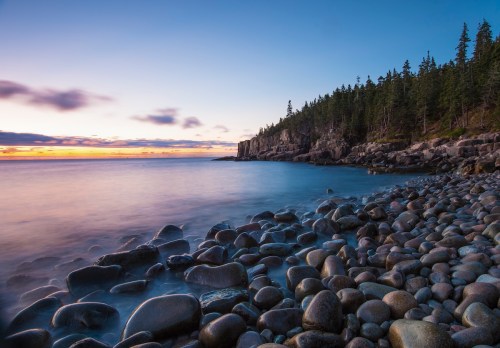 A serene coastal scene at sunset, featuring smooth stones, calm water, and a forested cliff in the background.