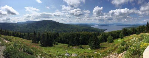 A panoramic view of lush green hills and a lake under a partly cloudy sky.