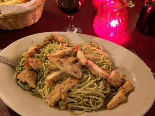 A plate of pasta with pesto sauce topped with grilled chicken, accompanied by a glass of red wine and a lit candle.