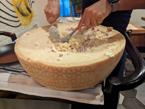A person mixes pasta in a large wheel of cheese, creating a creamy dish in a cozy restaurant setting.