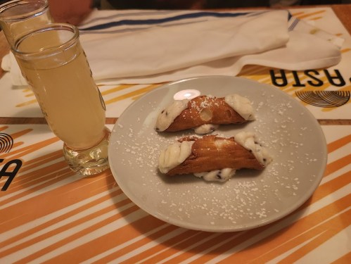 A plate with two cannoli filled with cream and dusted with powdered sugar, alongside a glass of lemon liqueur.