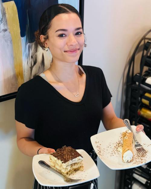 A smiling woman holds two plates of dessert: tiramisu and a cannoli, in a restaurant setting.