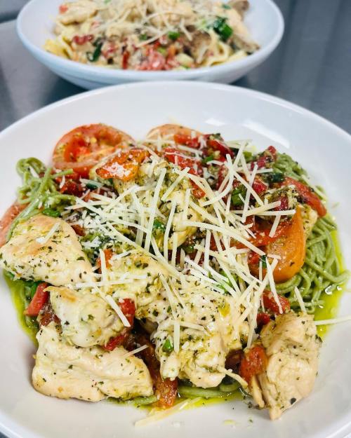A plate of green pasta topped with grilled chicken, tomatoes, herbs, and grated cheese, with another pasta dish in the background.