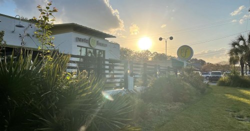 A restaurant exterior at sunset, featuring greenery and a sign, with a warm glow from the setting sun.