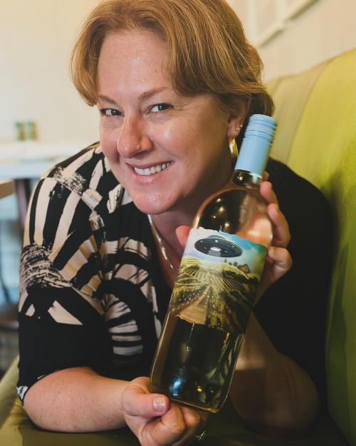 A smiling woman holds a bottle of wine featuring a unique label with a landscape and UFO design.