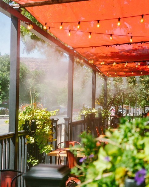 A vibrant outdoor patio with red canopies, string lights, and misting fans, surrounded by greenery and tables.