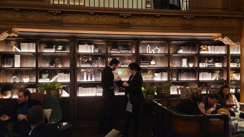 A cozy lounge with people socializing, a bookshelf filled with decor, and warm lighting creating an inviting atmosphere.