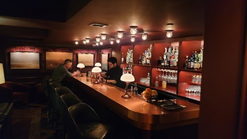 A dimly lit bar with two patrons, a bartender, and shelves stocked with various bottles and glasses.