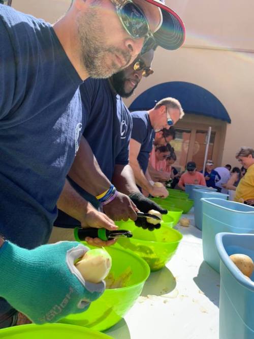 A group of people peeling potatoes at a community event, focused on their tasks with bowls in front of them.