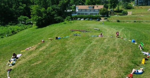 Aerial view of a grassy field with people arranged in a circle, surrounded by trees and a building in the background.