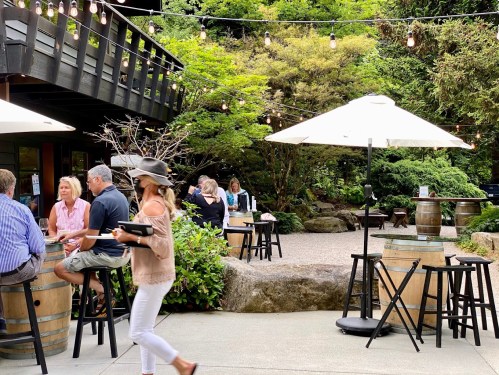 Outdoor dining area with string lights, tables, and patrons enjoying a meal in a lush garden setting.