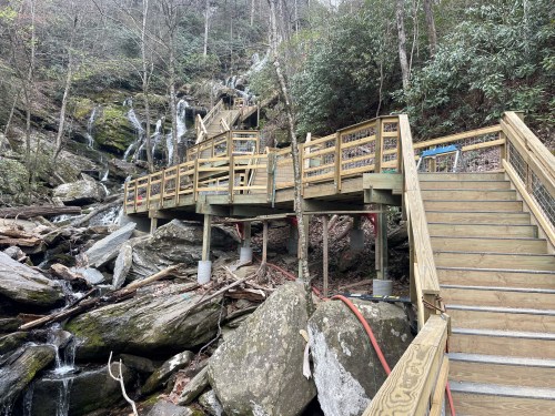 Wooden boardwalk and stairs alongside a rocky stream with waterfalls in a forested area.