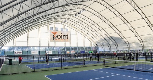 Indoor tennis facility with multiple courts, players in action, and a large "point" sign on the wall.