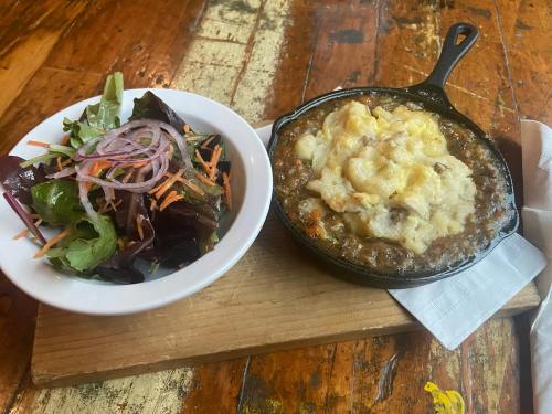 A cast iron skillet with baked dish beside a bowl of mixed salad with onions and carrots on a wooden board.