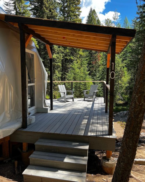A wooden deck with two chairs, surrounded by trees and nature, under a blue sky with clouds.