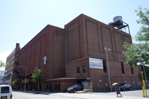 A large brick building with a cross on the side, featuring a water tower on the roof and trees nearby.