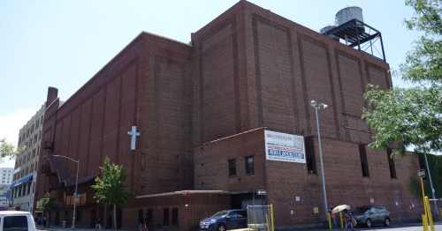 A large brick building with a cross on the side, featuring a water tower on the roof and trees nearby.