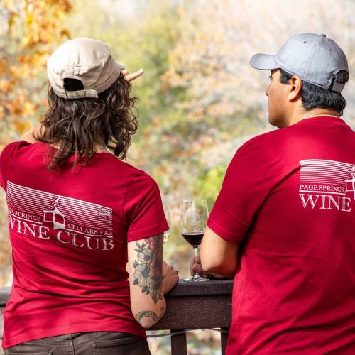 Two people in matching red shirts with "Wine Club" on the back, enjoying wine outdoors with autumn foliage in the background.
