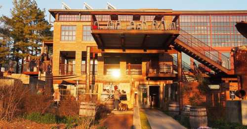 A modern brick building with large windows, outdoor seating, and a staircase, surrounded by greenery at sunset.