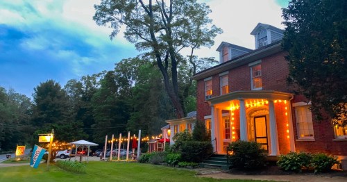 A charming brick building illuminated with string lights, surrounded by trees and a grassy area, under a twilight sky.