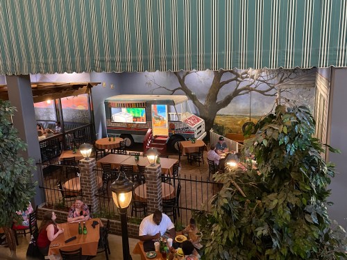 A cozy restaurant interior featuring a food truck mural, tables, and greenery, with diners enjoying their meals.