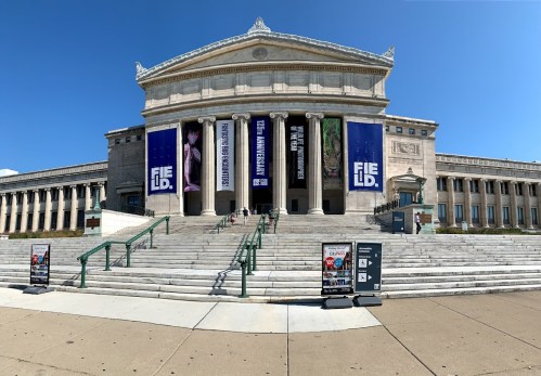 The exterior of a grand museum building with large banners and steps leading up to the entrance on a clear day.