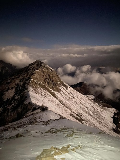 A snow-covered mountain peak under a night sky, surrounded by clouds and distant lights.