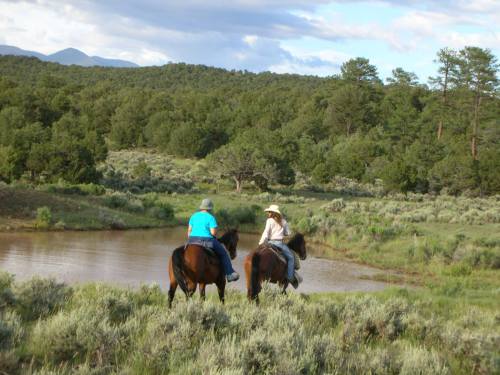 Two riders on horseback near a pond, surrounded by lush greenery and mountains in the background.