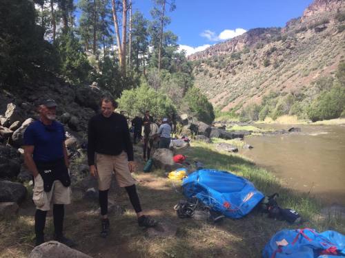 Two men stand by a riverbank, surrounded by trees and gear, with a group of people in the background preparing for an outing.