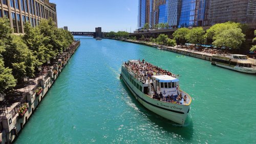 A crowded boat navigates a vibrant turquoise river surrounded by tall buildings and lush greenery on a sunny day.