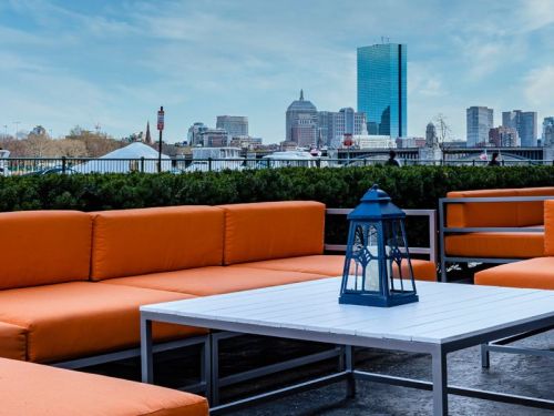A rooftop lounge area with orange seating and a blue lantern, overlooking a city skyline with tall buildings.