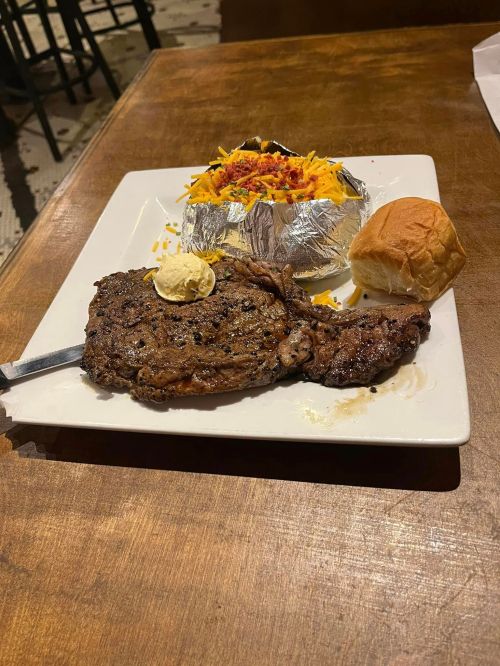 A plate with a grilled steak, a loaded baked potato topped with cheese and bacon, and a small dinner roll.