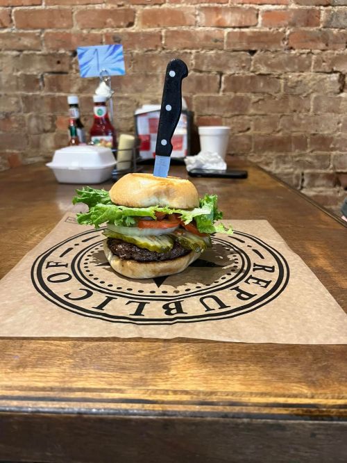 A burger with lettuce, tomato, and cheese, topped with a knife, on a branded paper on a wooden table.