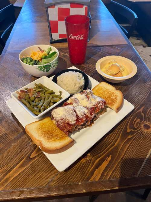 A plate with meatloaf, green beans, mashed potatoes, toast, a salad, and a dessert, with a red drink on the table.