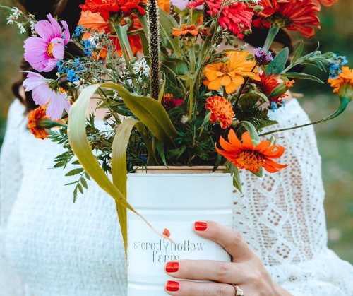 A person holding a white vase filled with colorful flowers, wearing a lace top and showing off red nail polish.