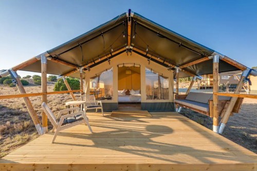 A spacious glamping tent with a wooden deck, featuring large windows and a cozy interior, set in a natural landscape.